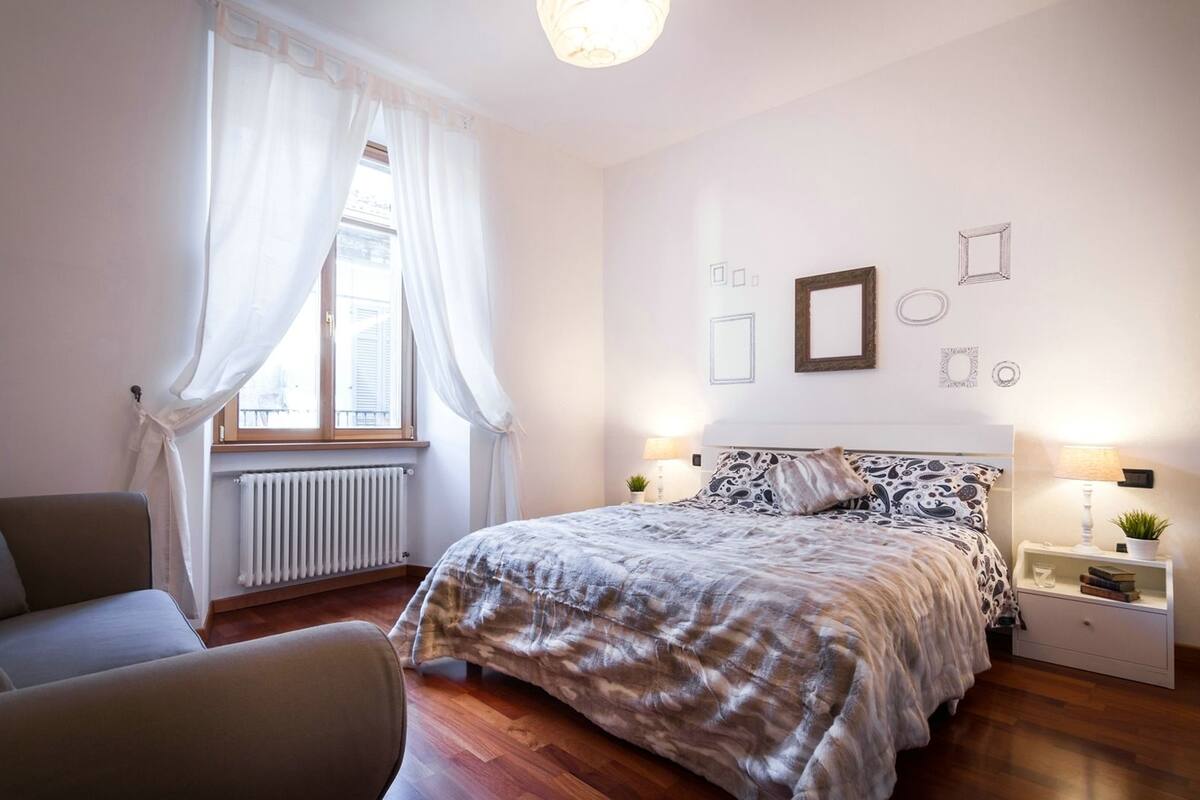 Quarto com cama, uma cadeira, mesa de cabeceira com luminária, radiador e janela com cortinas.