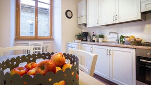 A kitchen with white cabinets, a window with blinds, a clock on the wall, and a basket of apples on the table. - La Contrada del Pittore (Como)
