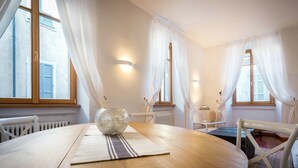 A round dining table with a glass bowl centerpiece, white curtains, and a view of a building through the windows. - La Contrada del Pittore (Como)