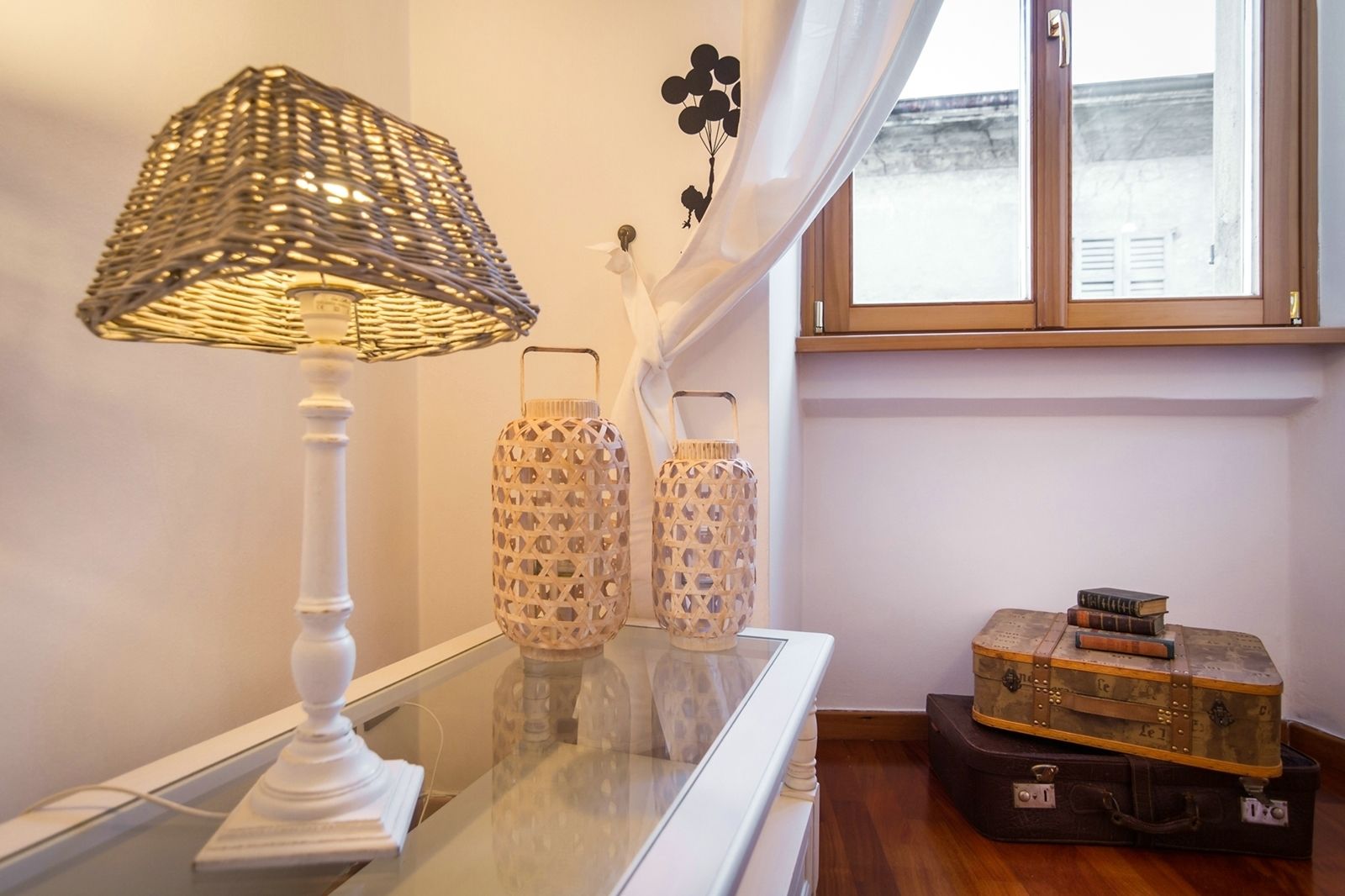 A room with a lamp, two decorative lanterns, a vase with flowers, and a vintage suitcase on a table.