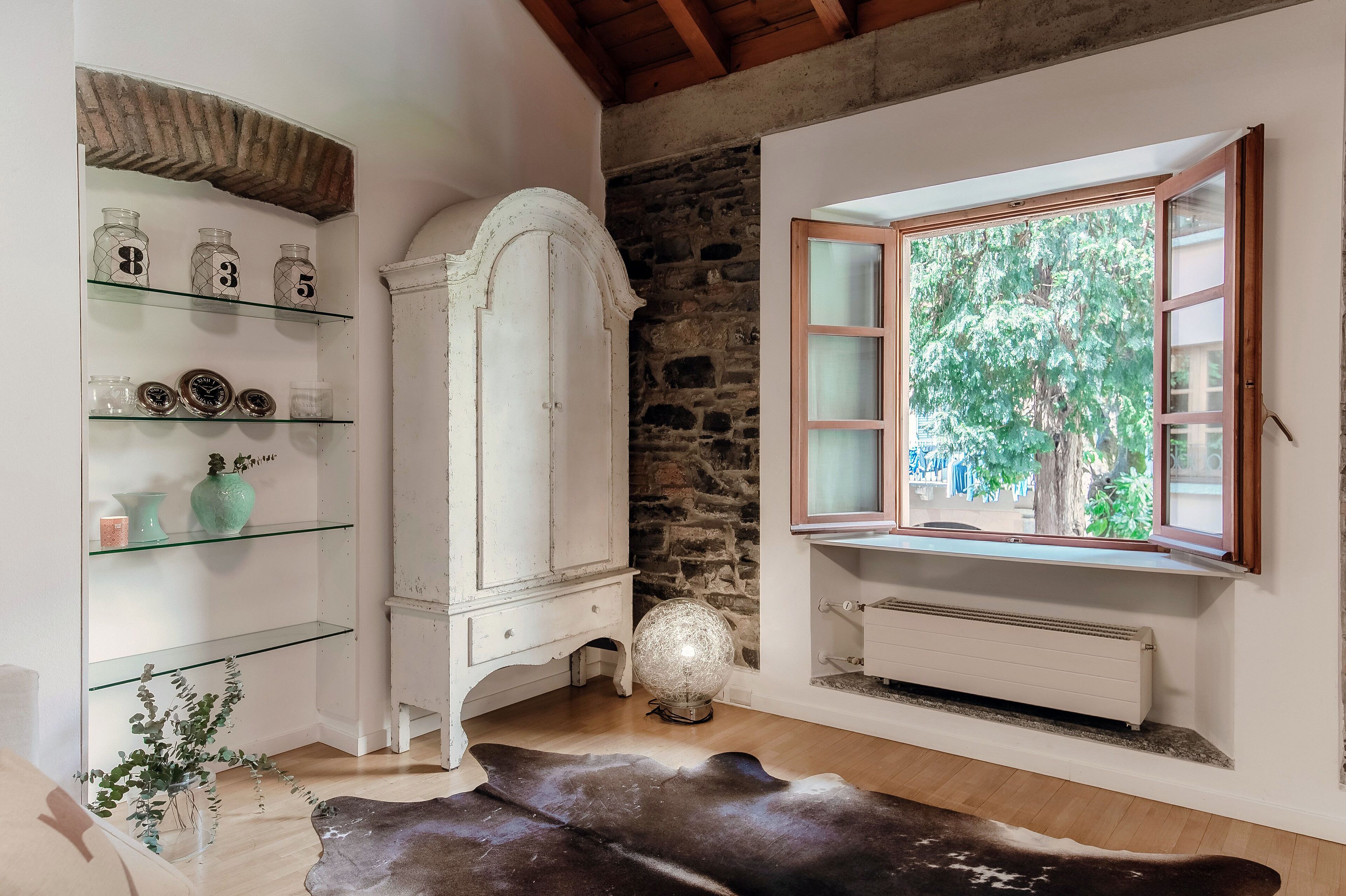 A room with a white cabinet with glass shelves, a stone wall, a radiator, and a window with a view of trees.