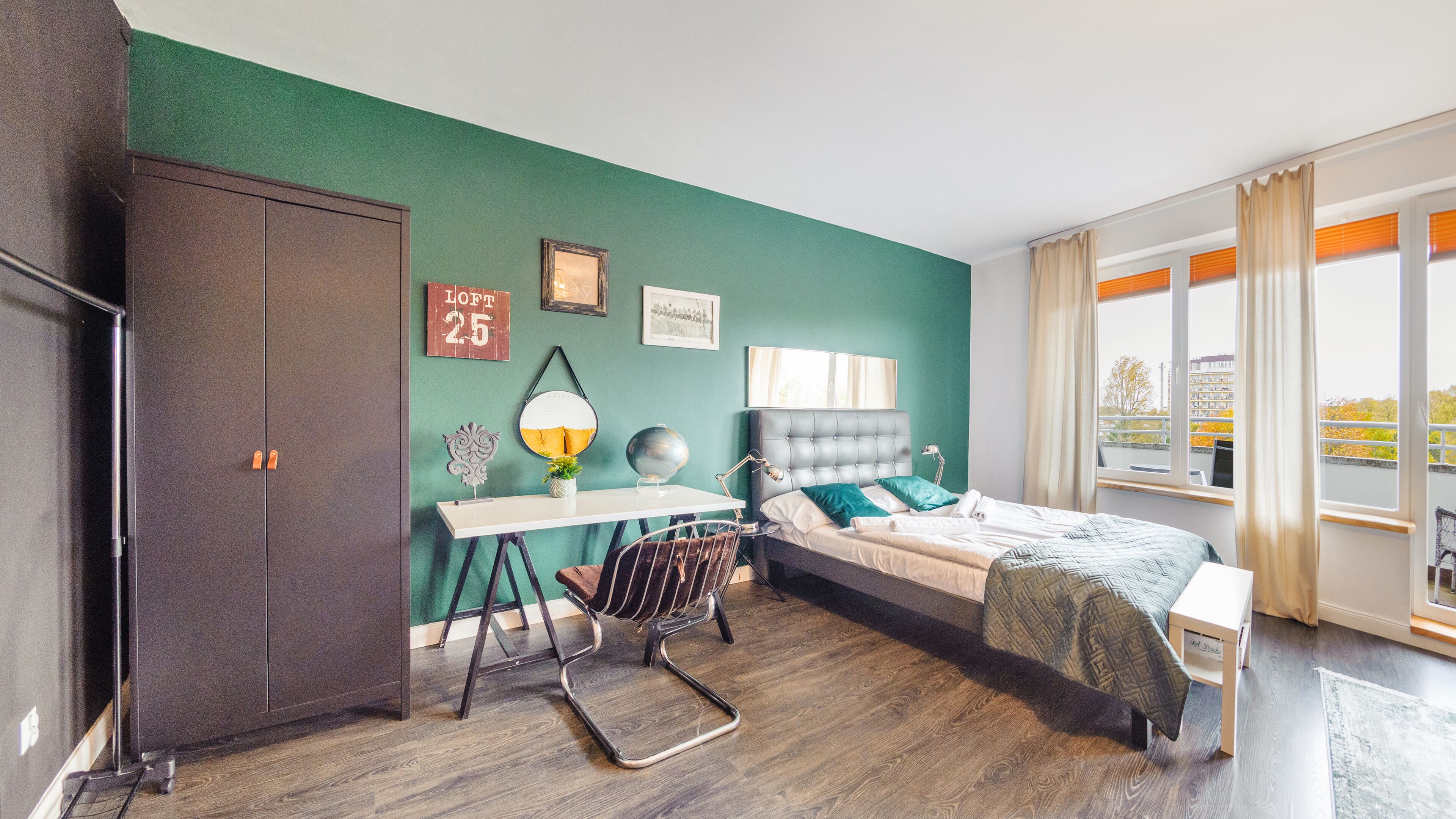 A modern bedroom with a green wall, a bed with a grey headboard, a desk with a chair, and a wardrobe.