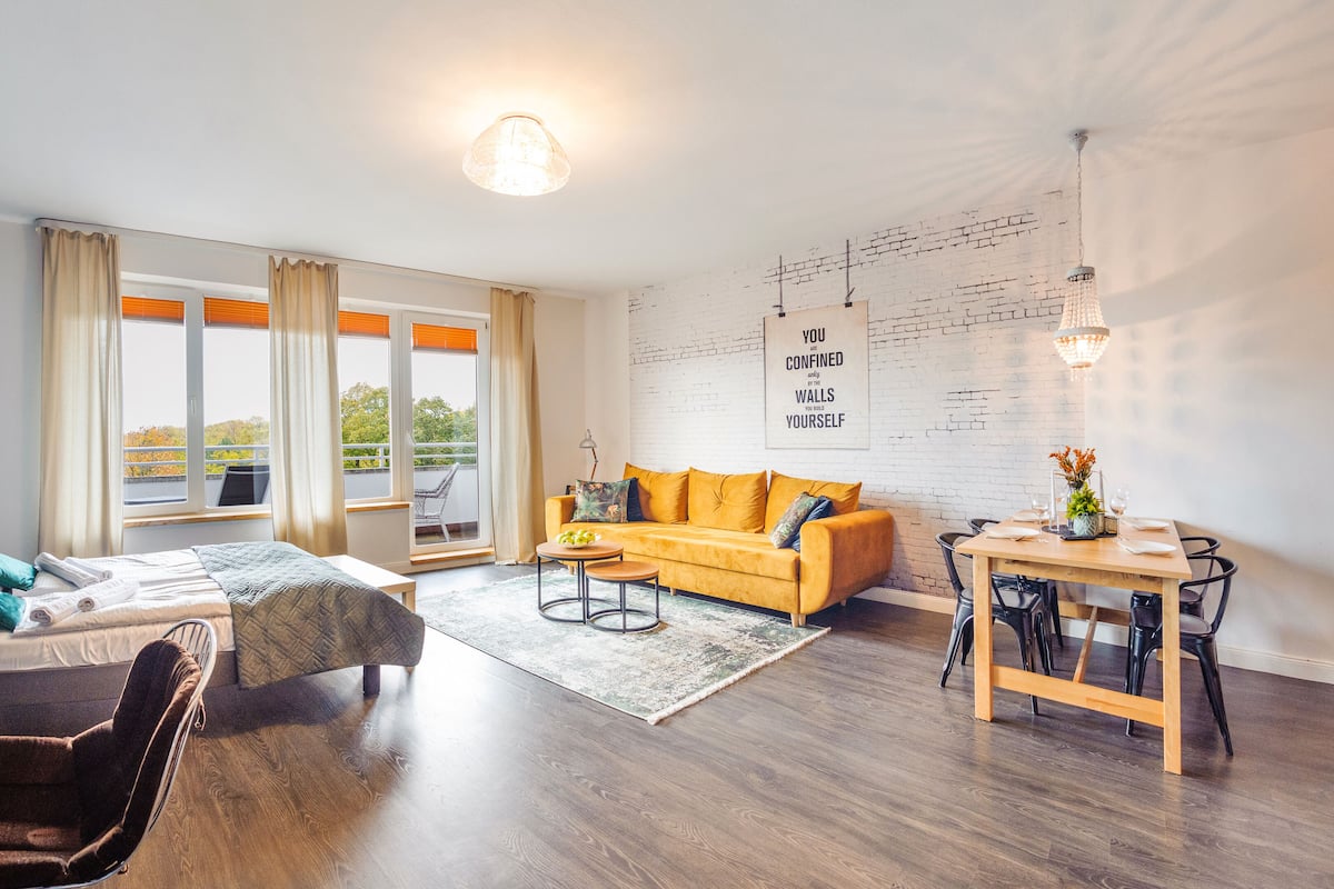 A modern living room with a sofa, a dining table, and a large window offering a view of the outdoors.