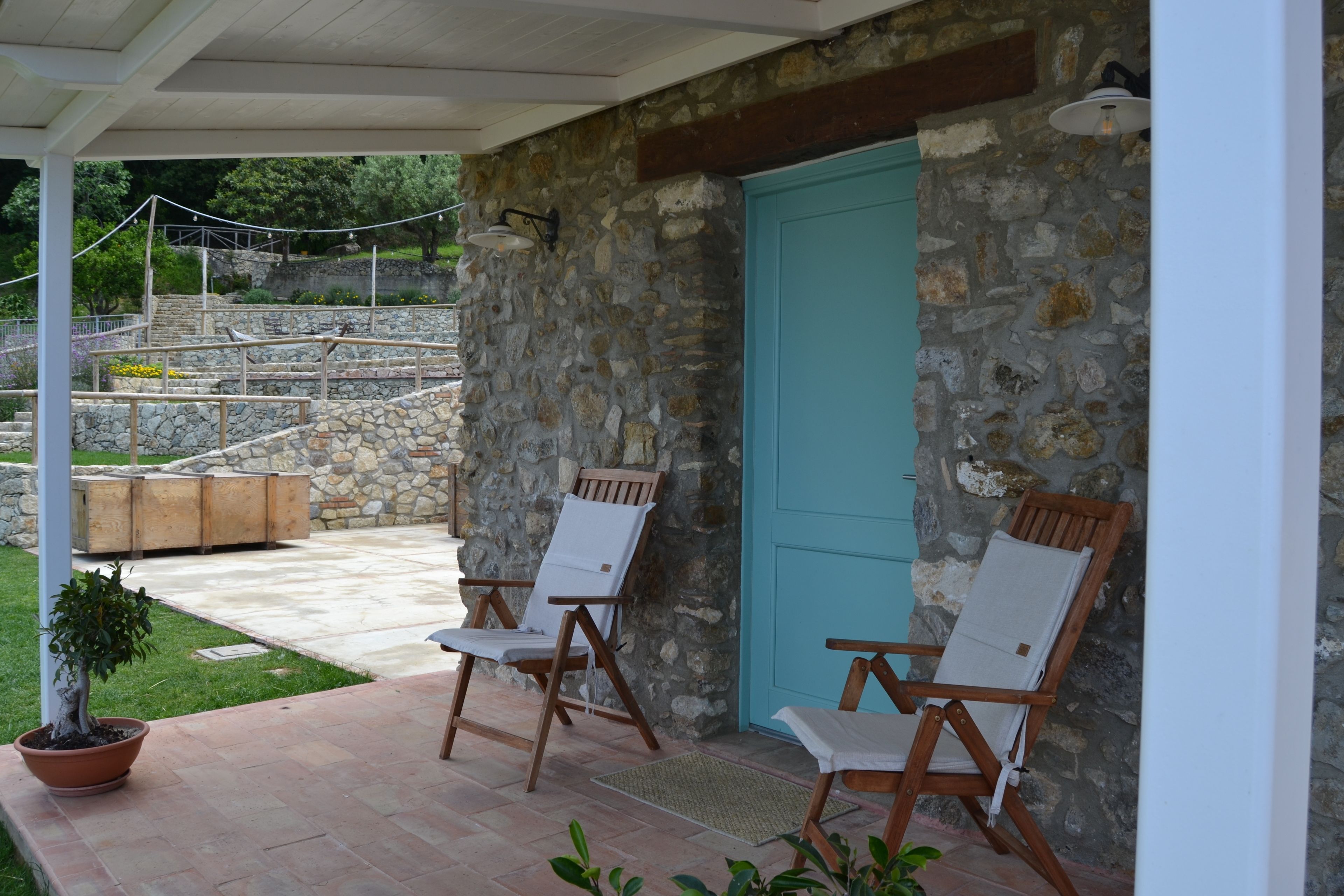 Une terrasse couverte avec des murs en pierre, deux fauteuils de jardin en bois, une plante en pot et une porte bleue.