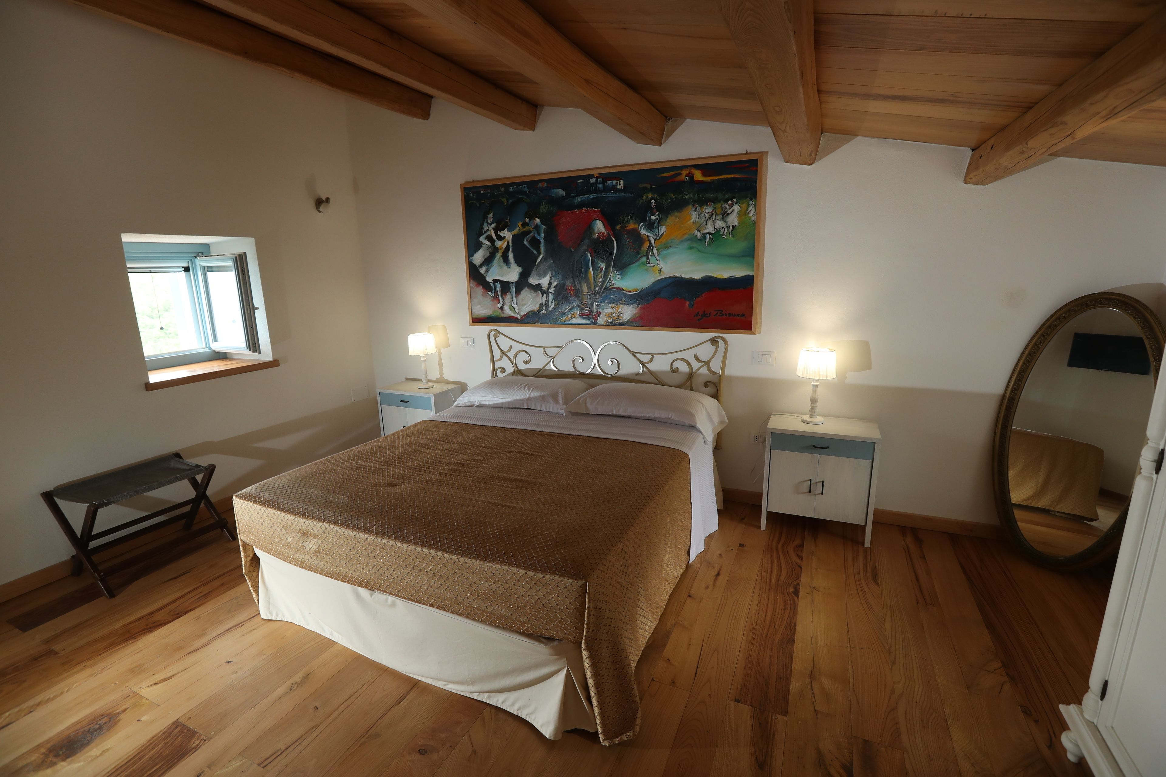 A bedroom with a large bed, wooden flooring, a painting above the bed, a window, bedside tables with lamps, and a mirror.