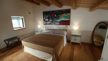 A bedroom with a large bed, wooden flooring, a painting above the bed, a window, bedside tables with lamps, and a mirror.