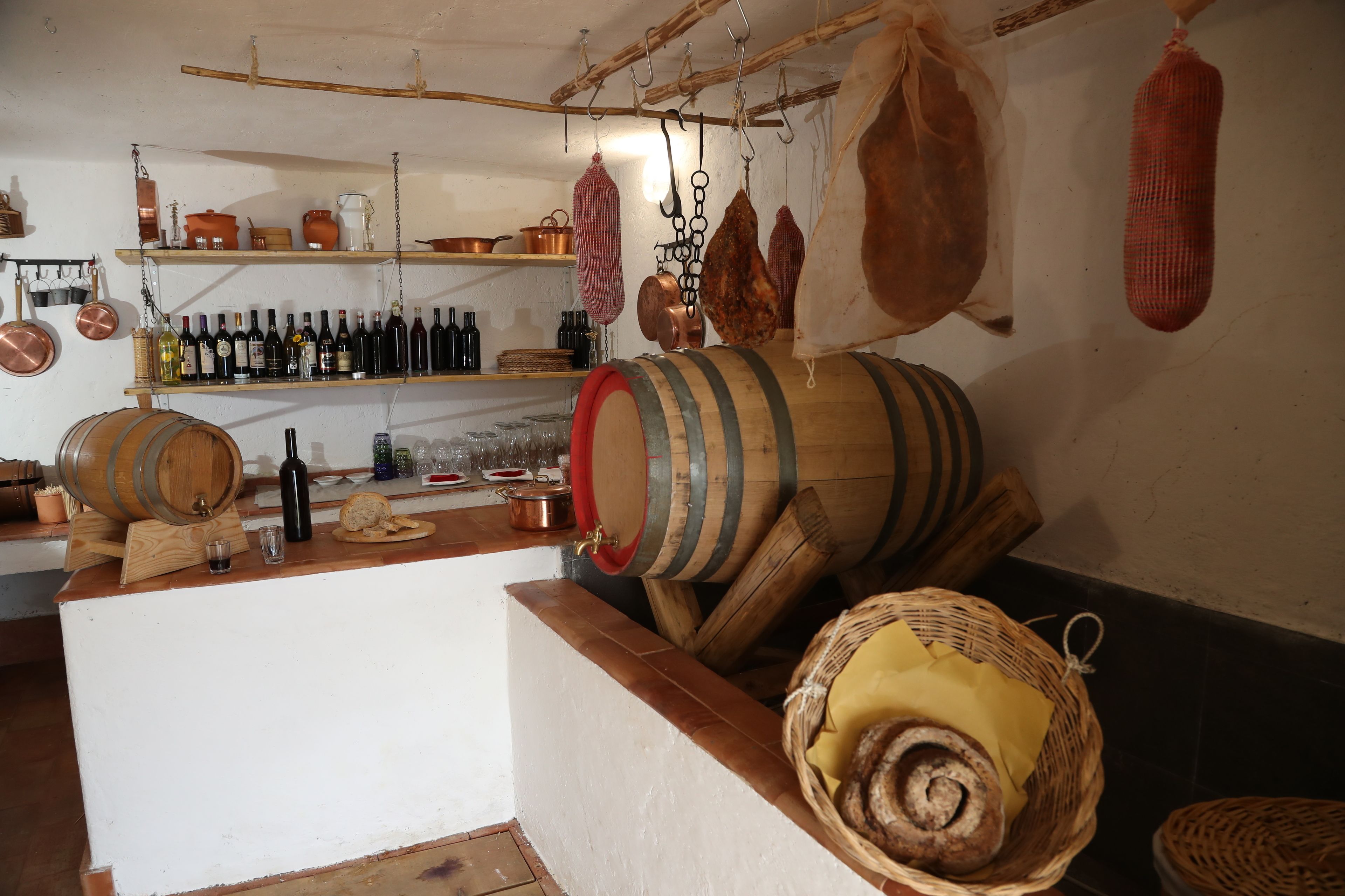 Cocina rústica con barriles de madera, estantes con botellas y carne colgando.