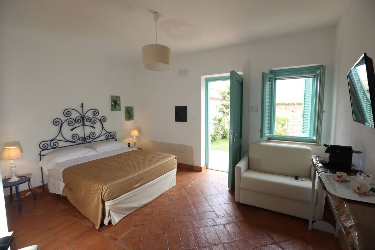 Une chambre à coucher avec un lit, une table de chevet, une lampe et une vue sur l’extérieur, grâce à une porte ouverte.