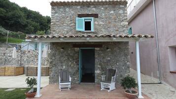 A stone building with a small porch, two chairs, and two potted plants.