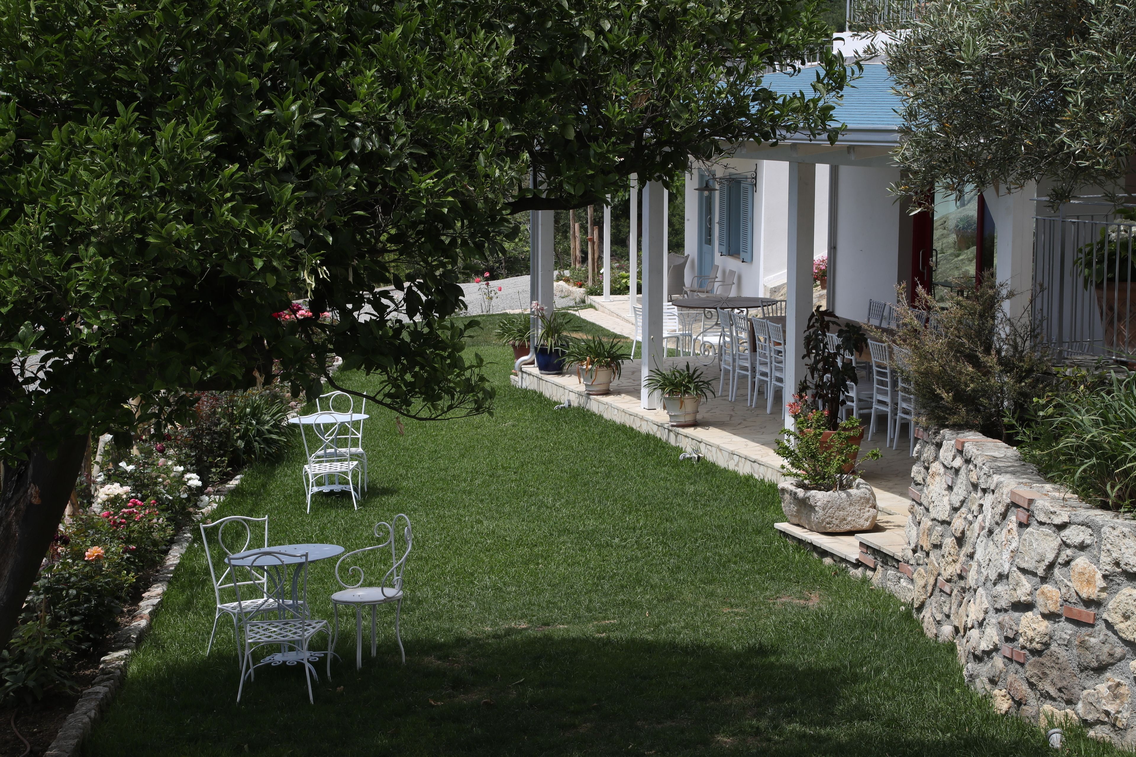 Une terrasse de jardin avec des chaises et des tables en métal blanc, entourée d’un mur de pierre et d’une végétation luxuriante.