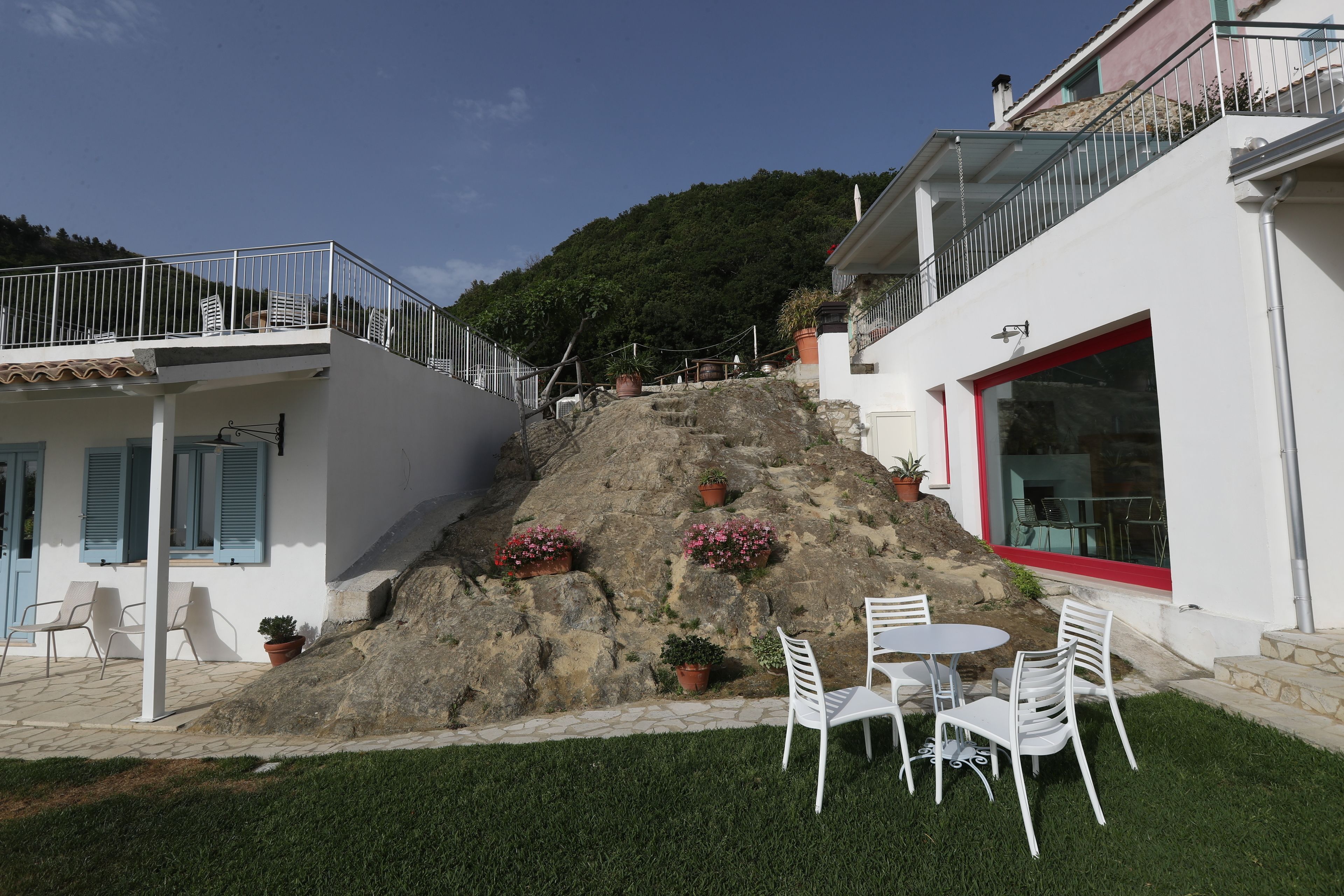 Un edificio con fachada blanca, una ventana con marco rojo y un balcón con barandilla metálica. Hay plantas en macetas en la fachada y un área con césped, sillas blancas y una mesa.