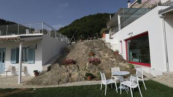 Un edificio con fachada blanca, una ventana con marco rojo y un balcón con barandilla metálica. Hay plantas en macetas en la fachada y un área con césped, sillas blancas y una mesa.