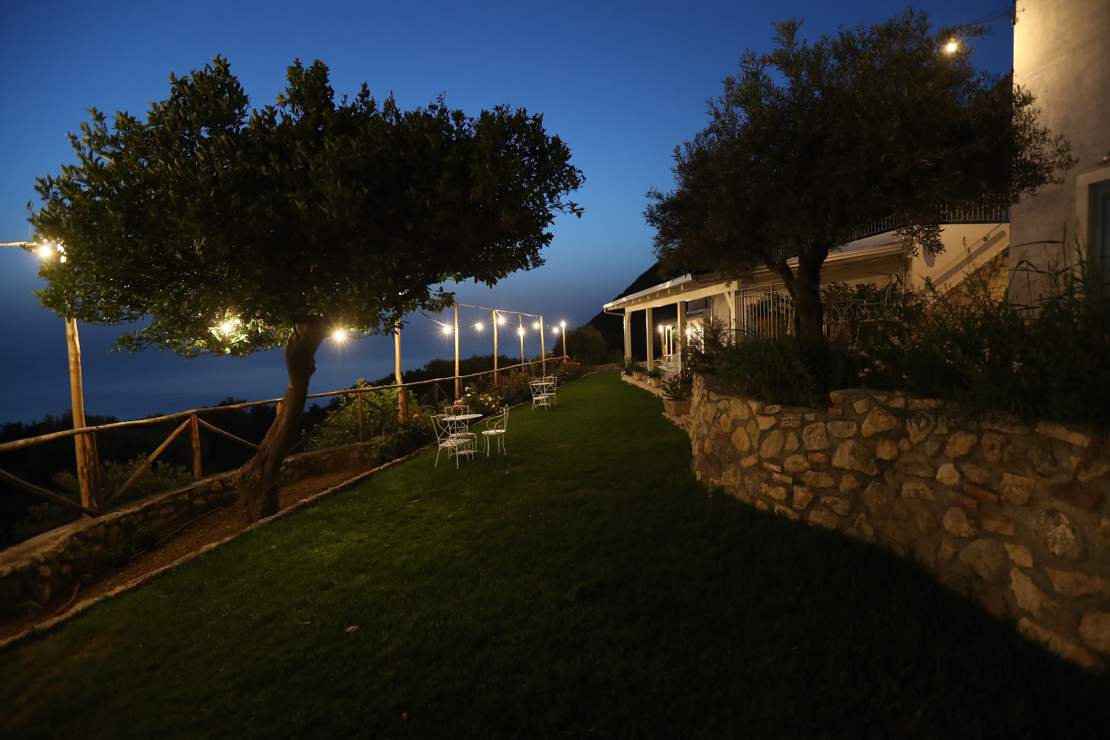A well-lit outdoor patio area with stone walls, a grassy lawn, and tables and chairs set up for dining.