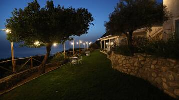A well-lit outdoor patio area with stone walls, a grassy lawn, and tables and chairs set up for dining.