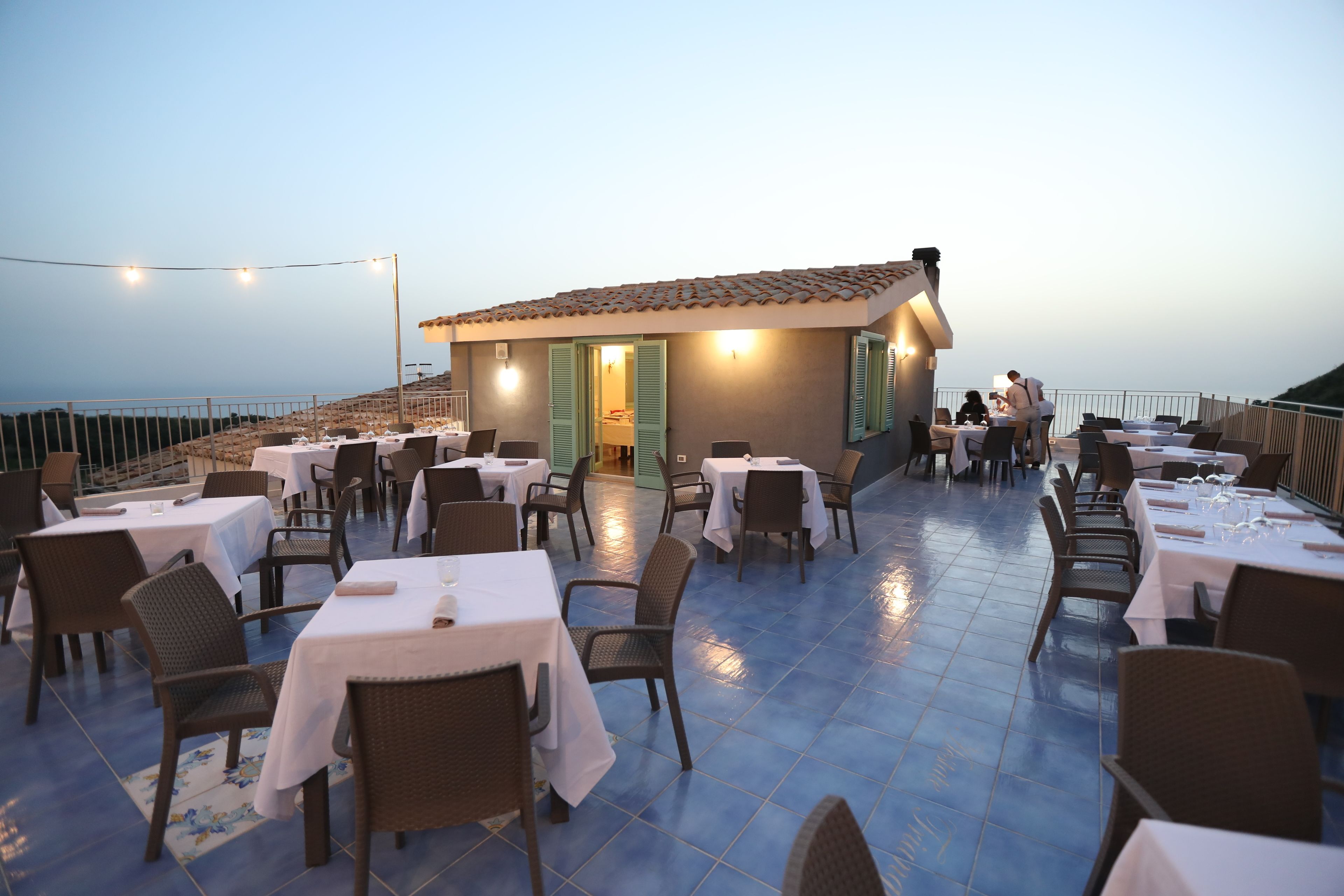 An outdoor dining area with tables and chairs, a building with green shutters, and a view of the sea.