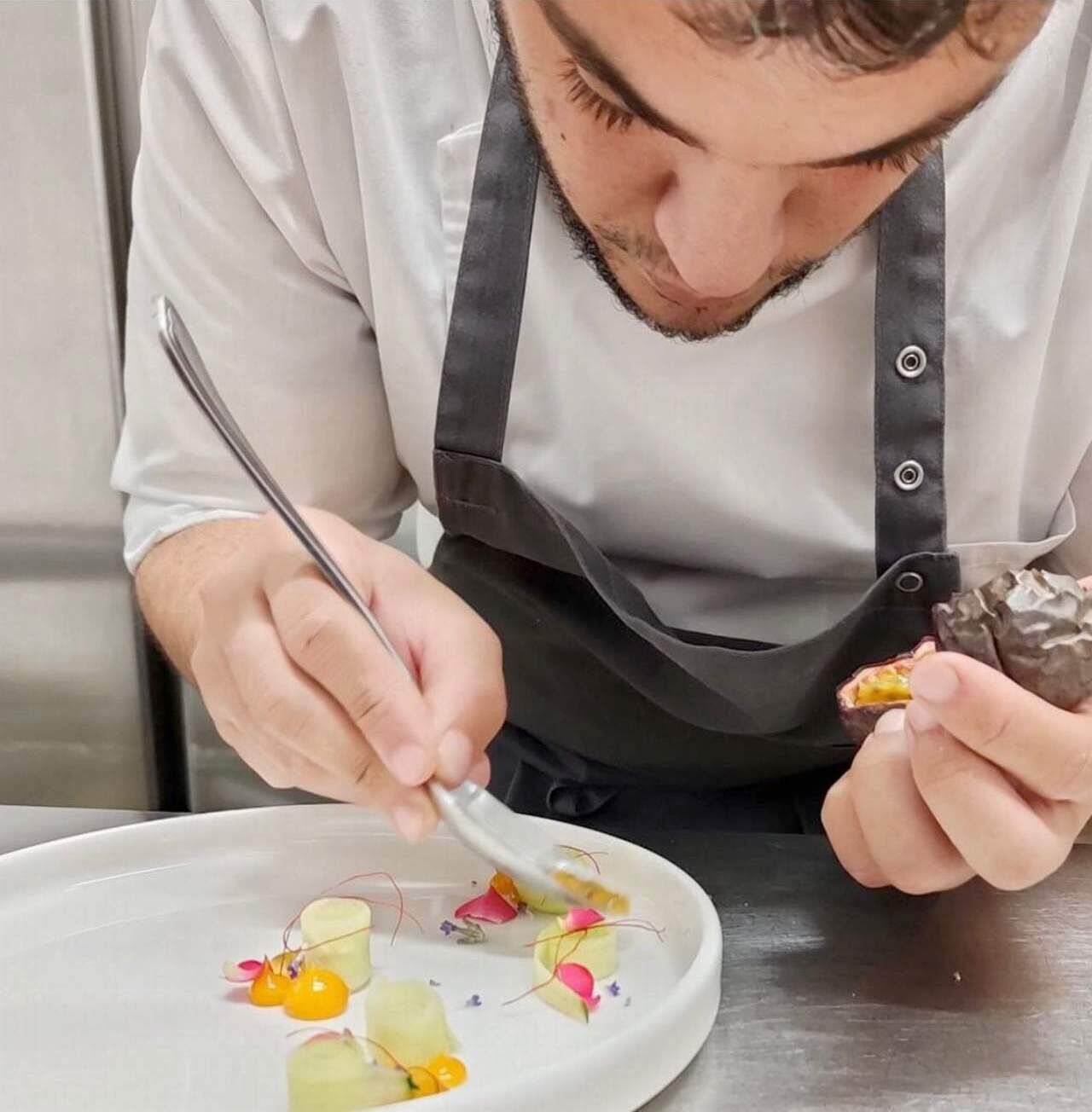 Une personne vêtue d’une chemise blanche et d’un tablier noir prépare un plat à l’aide d’une cuillère.