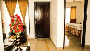 A hotel room with a bed, a desk with a vase of flowers, and a door leading to another room. - Van Durga Villas & Suites Katra (Reasi)