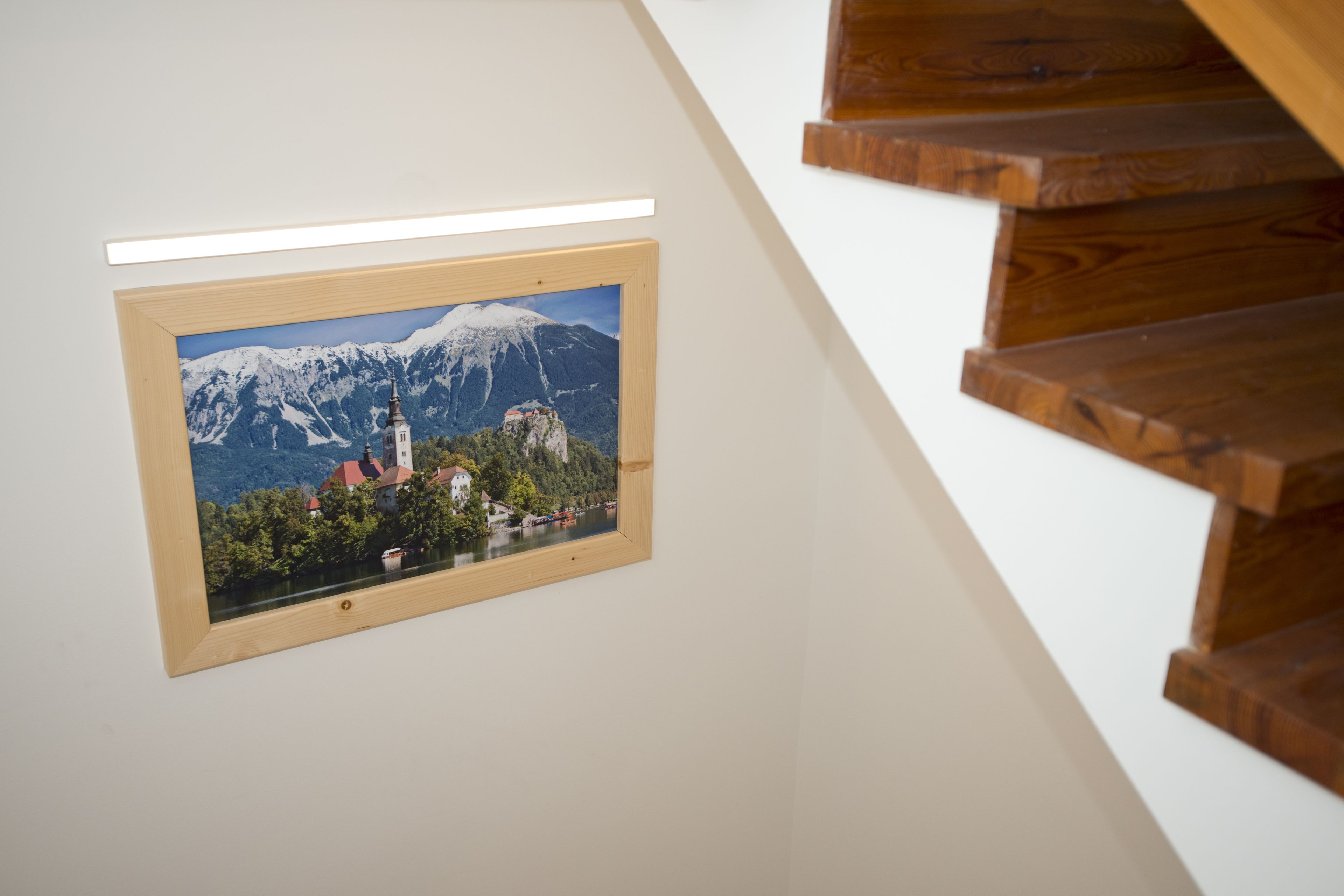 A framed photograph of a mountain landscape with a church and lake, mounted on a white wall next to a wooden staircase.