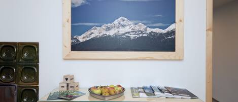 A desk with a framed mountain landscape, a bowl of apples, and some magazines.