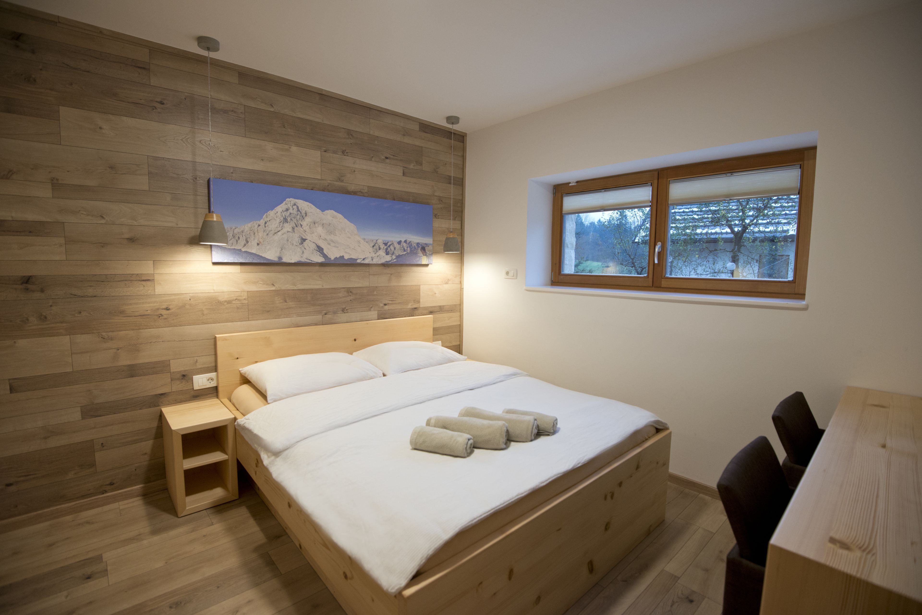 A bedroom with a wooden bed, a nightstand, a window with a view of trees, and a painting of mountains on the wall.