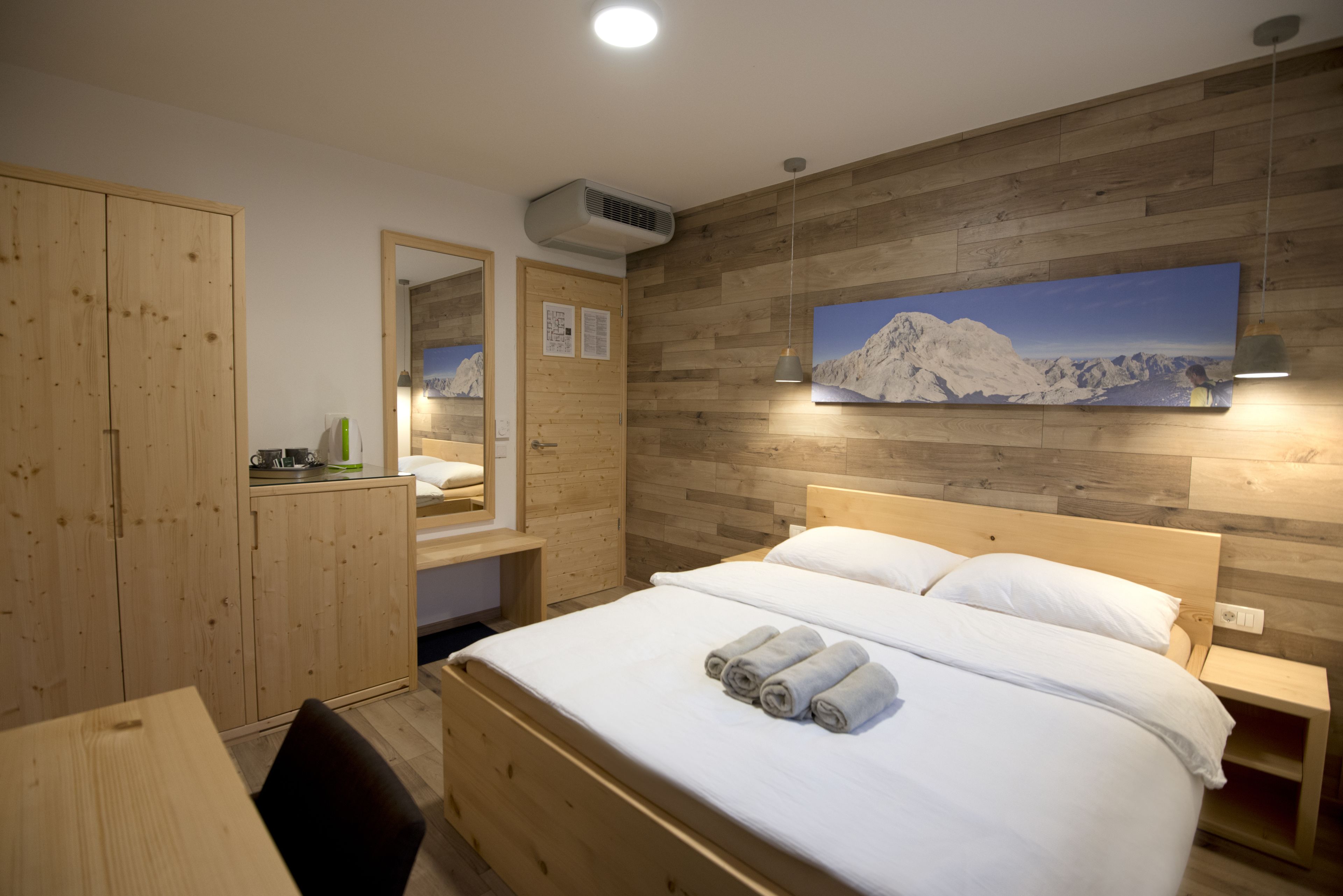 A hotel room with a wooden bed, a wooden wardrobe, a mirror, and a mountain painting on the wall.