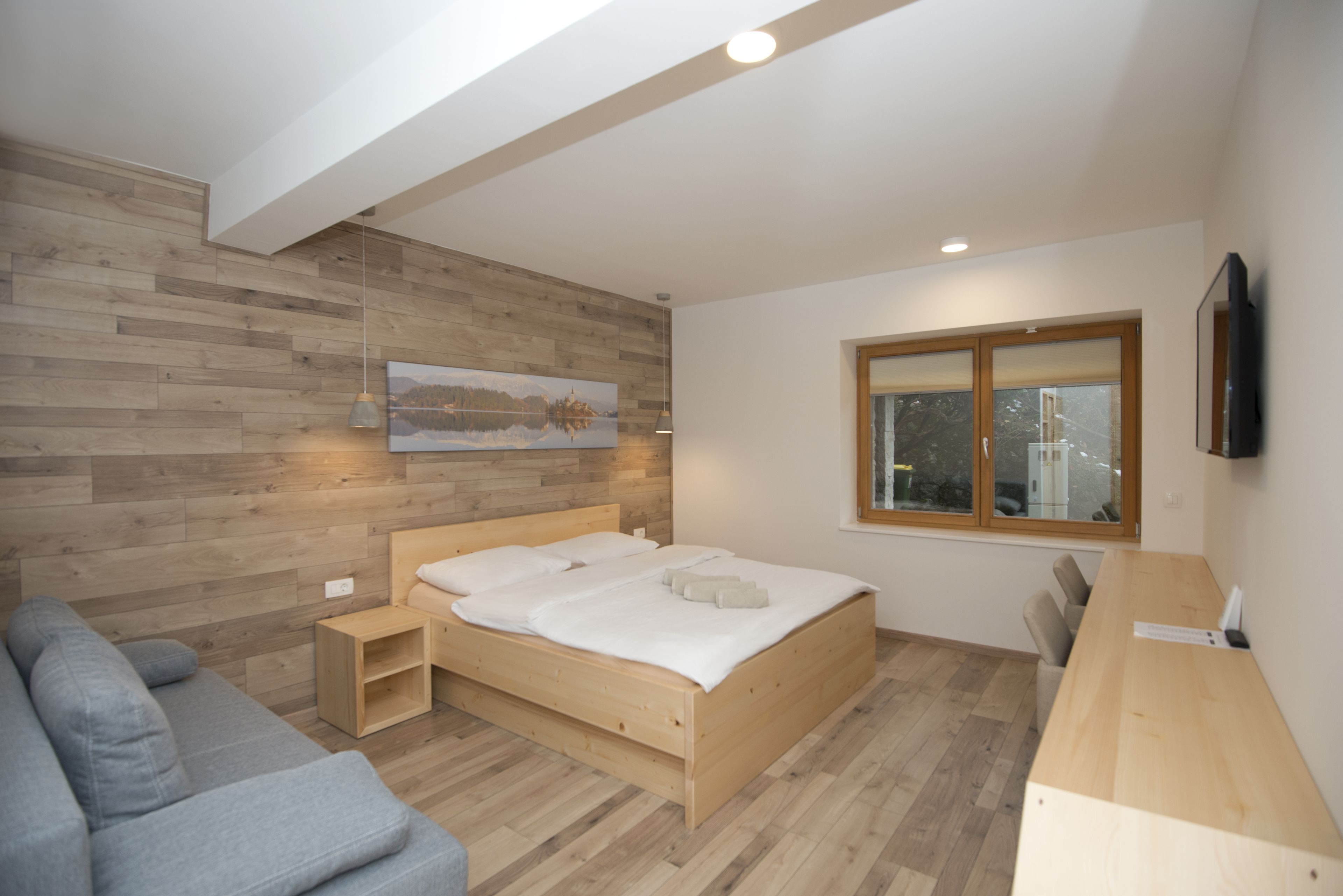 A modern bedroom with a wooden bed, a gray sofa, a wooden desk, and a window with a view of the city.