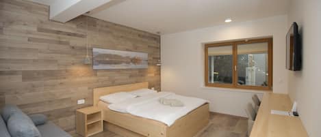 Un dormitorio moderno con cama de madera, sofá gris, escritorio de madera y ventana con vista a la ciudad.