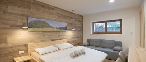Un dormitorio con cabecera de madera, cama con ropa de cama blanca, un sofá gris y una ventana con vistas a árboles.