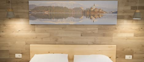 A wooden-paneled wall with a framed landscape painting, a bed with white linens, and two pendant lights.