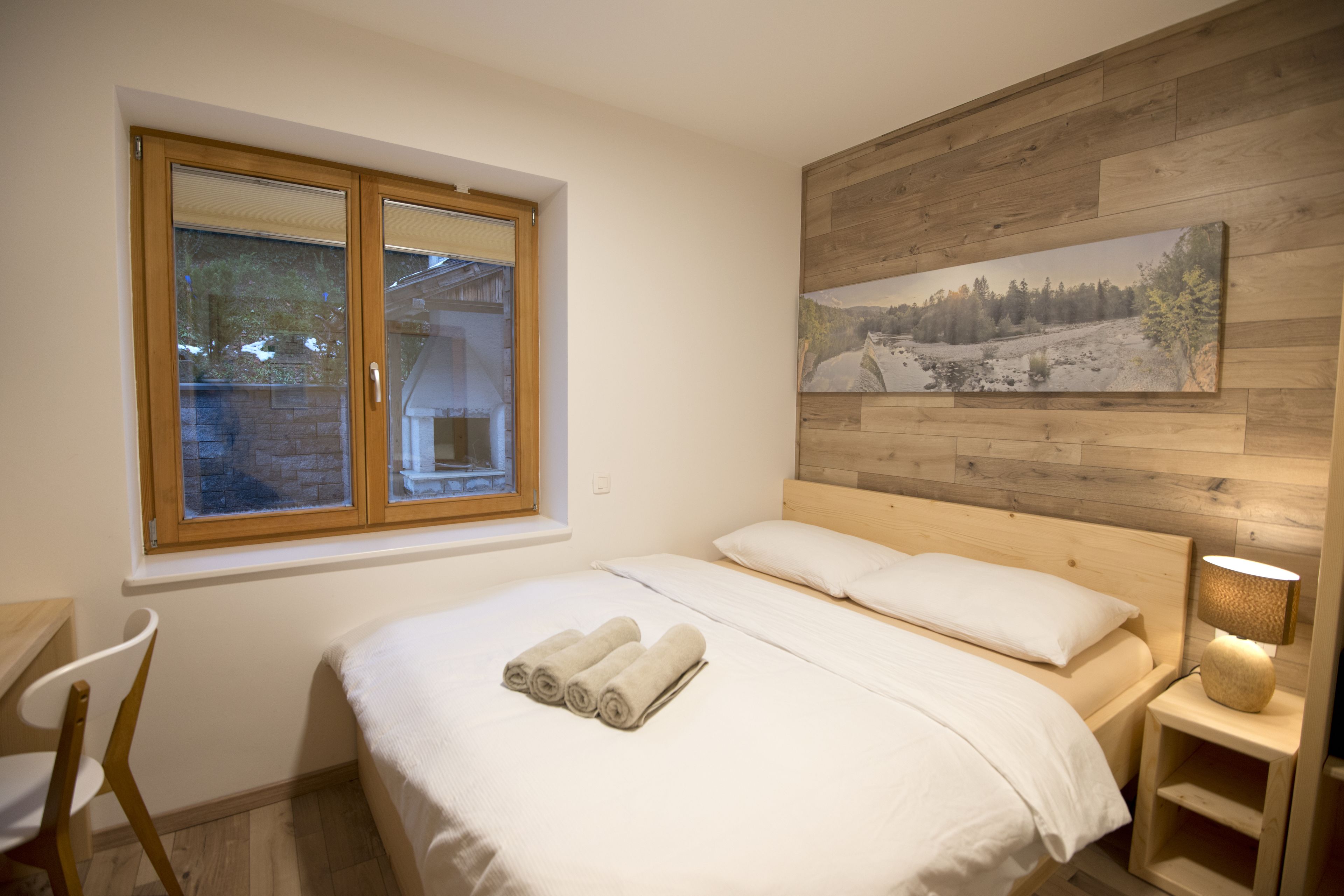 A bedroom with a wooden headboard, a large window, a painting on the wall, and a bedside table with a lamp.