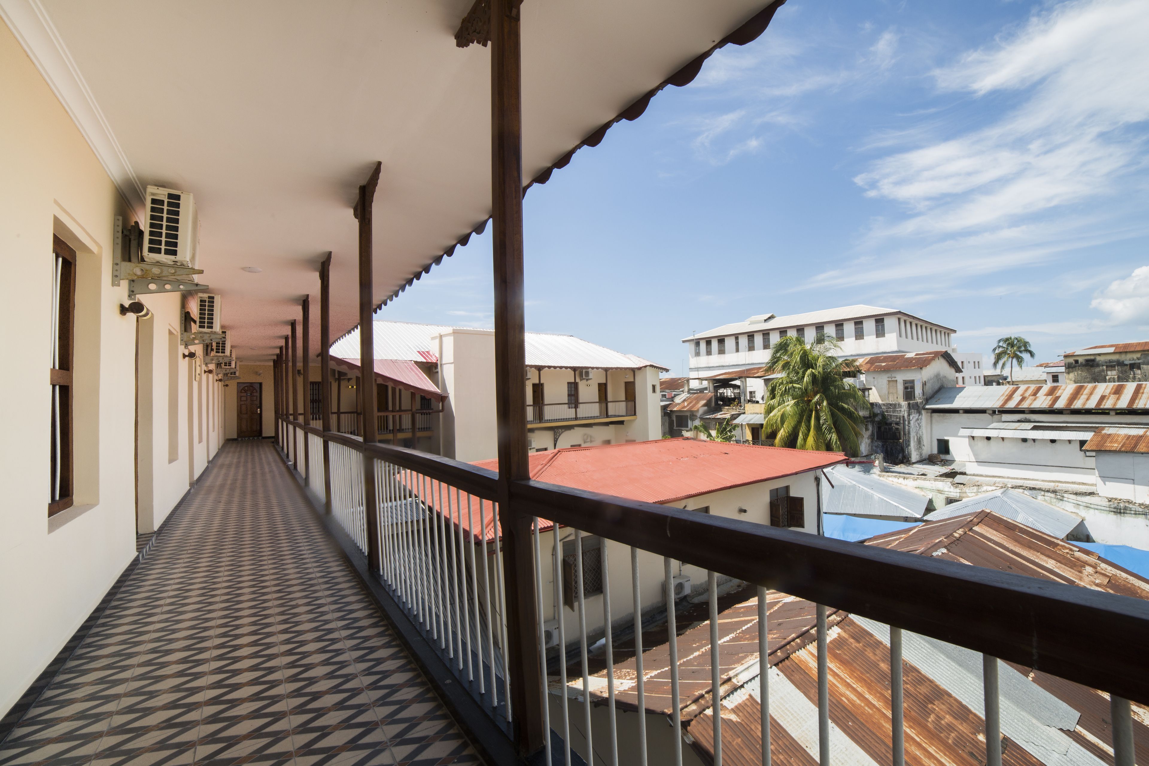 Un couloir au sol décoré de motifs, un balcon avec une balustrade, et des bâtiments en arrière-plan.