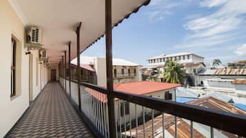 Un couloir au sol décoré de motifs, un balcon avec une balustrade, et des bâtiments en arrière-plan.