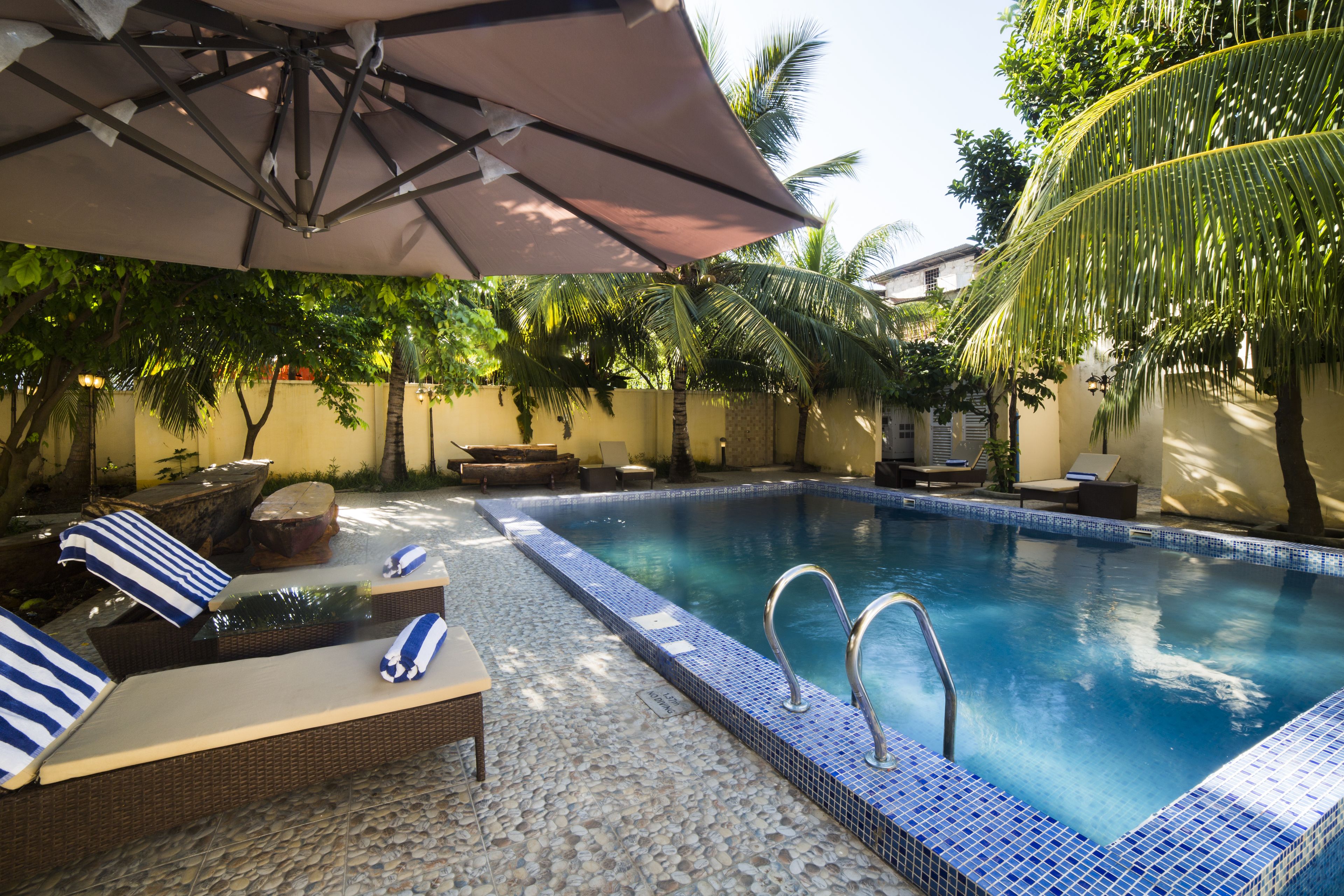 Ein Pool mit Liegestühlen und Sonnenschirm, umgeben von Palmen und einer gefliesten Terrasse.