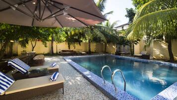 Ein Pool mit Liegestühlen und Sonnenschirm, umgeben von Palmen und einer gefliesten Terrasse.