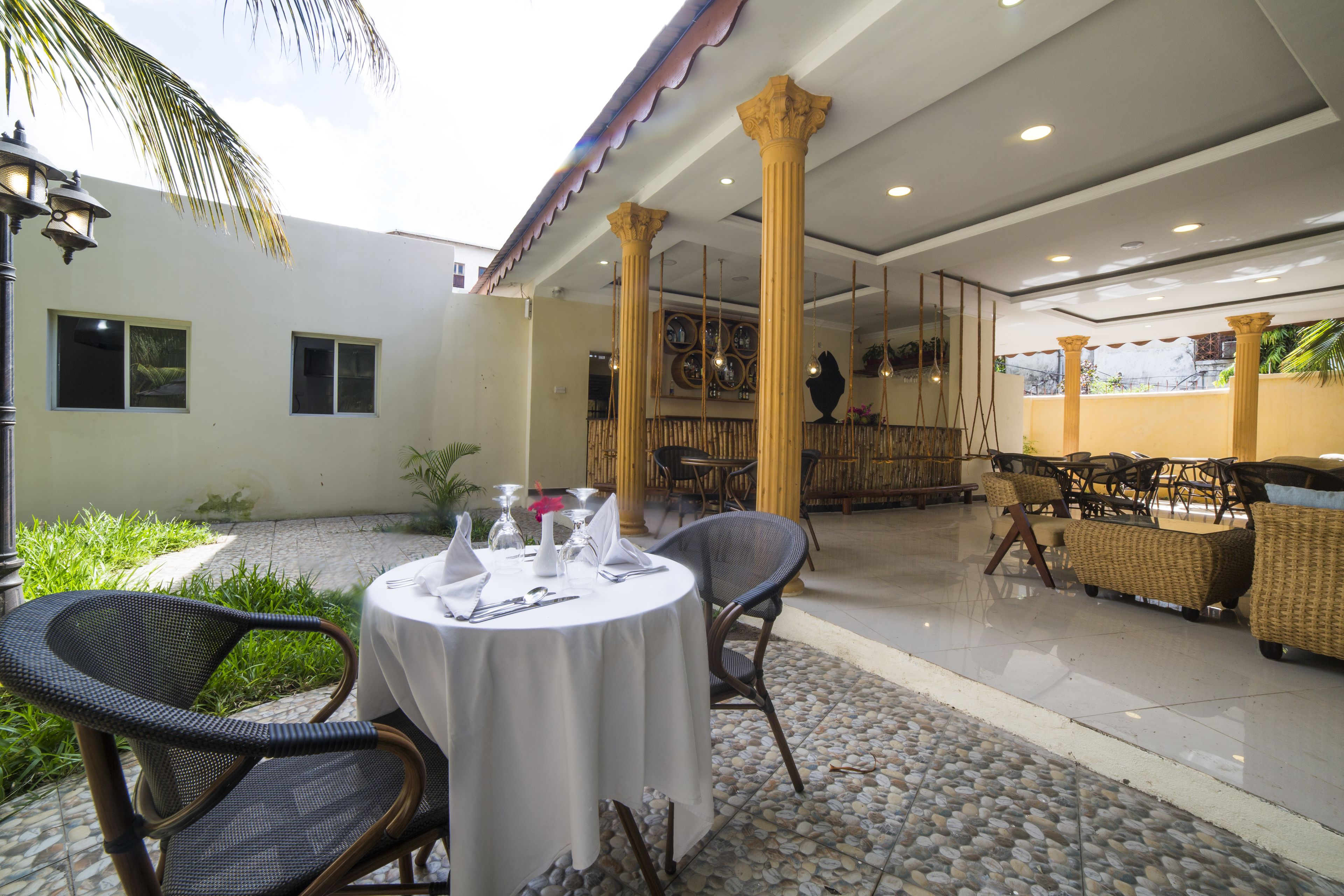 A covered outdoor dining area with round tables, wicker chairs, and a stone-tiled floor.