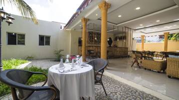 A covered outdoor dining area with round tables, wicker chairs, and a stone-tiled floor.