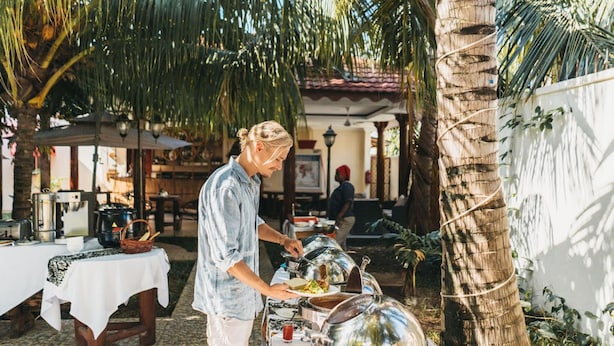 Une personne servant des plats dans un buffet en plein air, avec des présentoirs couverts, des palmiers et une terrasse carrelée.