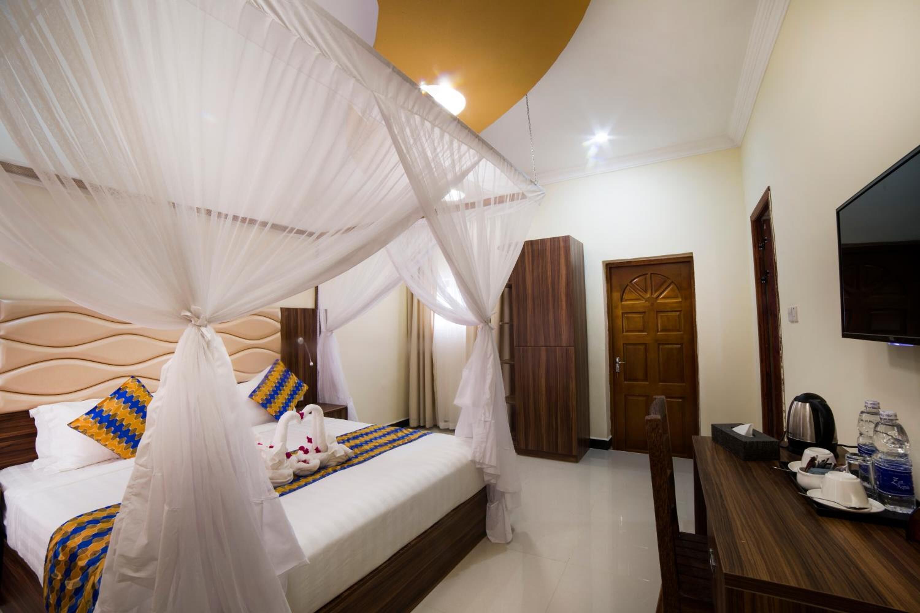 A hotel room with a bed, a TV, a wooden wardrobe, and a desk with a kettle and water bottles.