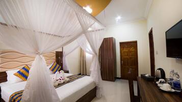 A hotel room with a bed, a TV, a wooden wardrobe, and a desk with a kettle and water bottles.