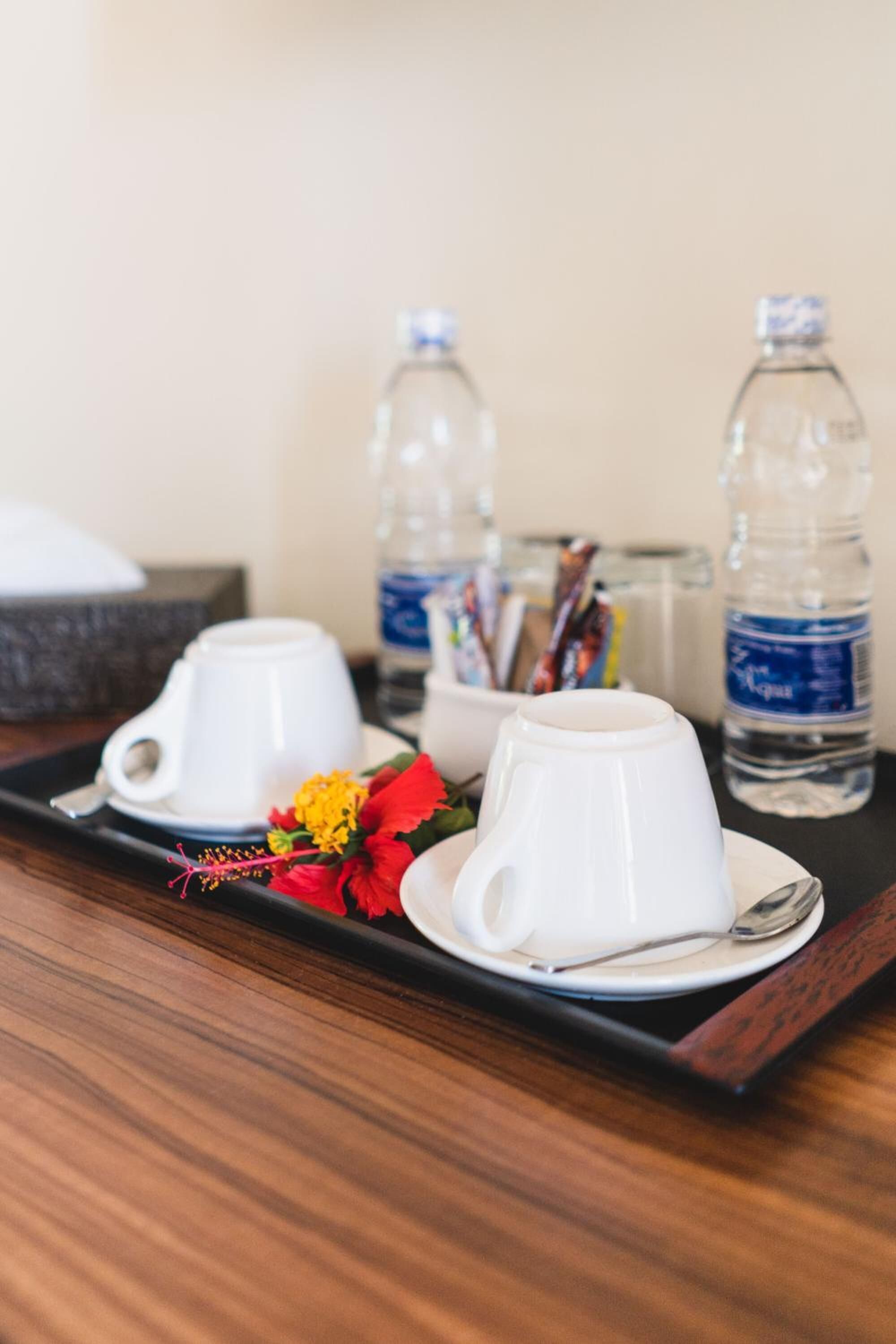 Una bandeja con dos tazas blancas y un cuenco pequeño, acompañada de una botella de agua y una flor.