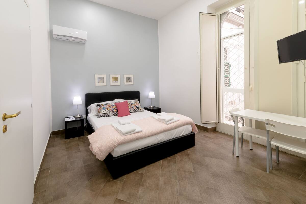 A bedroom with a bed, bedside tables, a desk, and a large window.