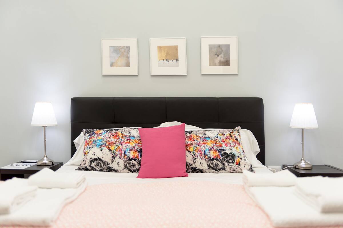 A neatly made bed with a black headboard, two bedside lamps, and three framed artworks on the wall.