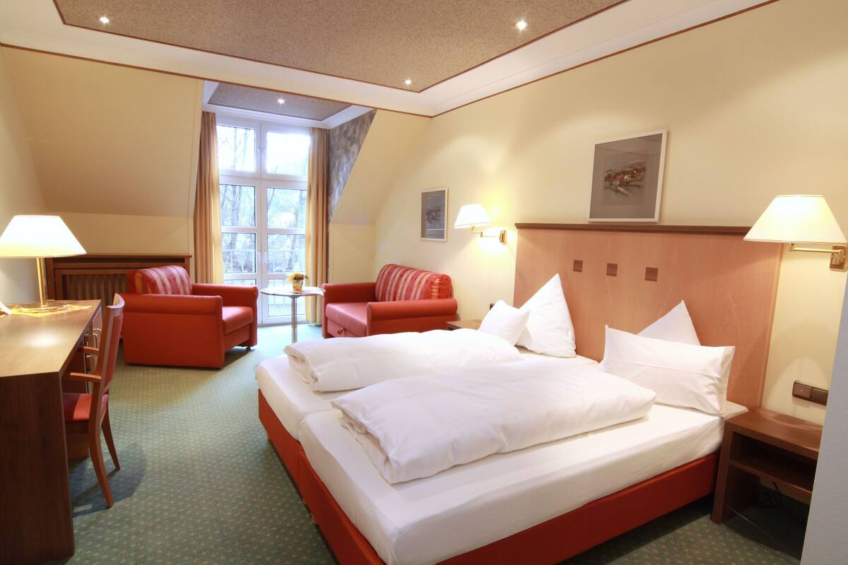 Quarto de hotel com uma cama grande, dois sofás vermelhos, uma escrivaninha de madeira e uma janela com cortinas.