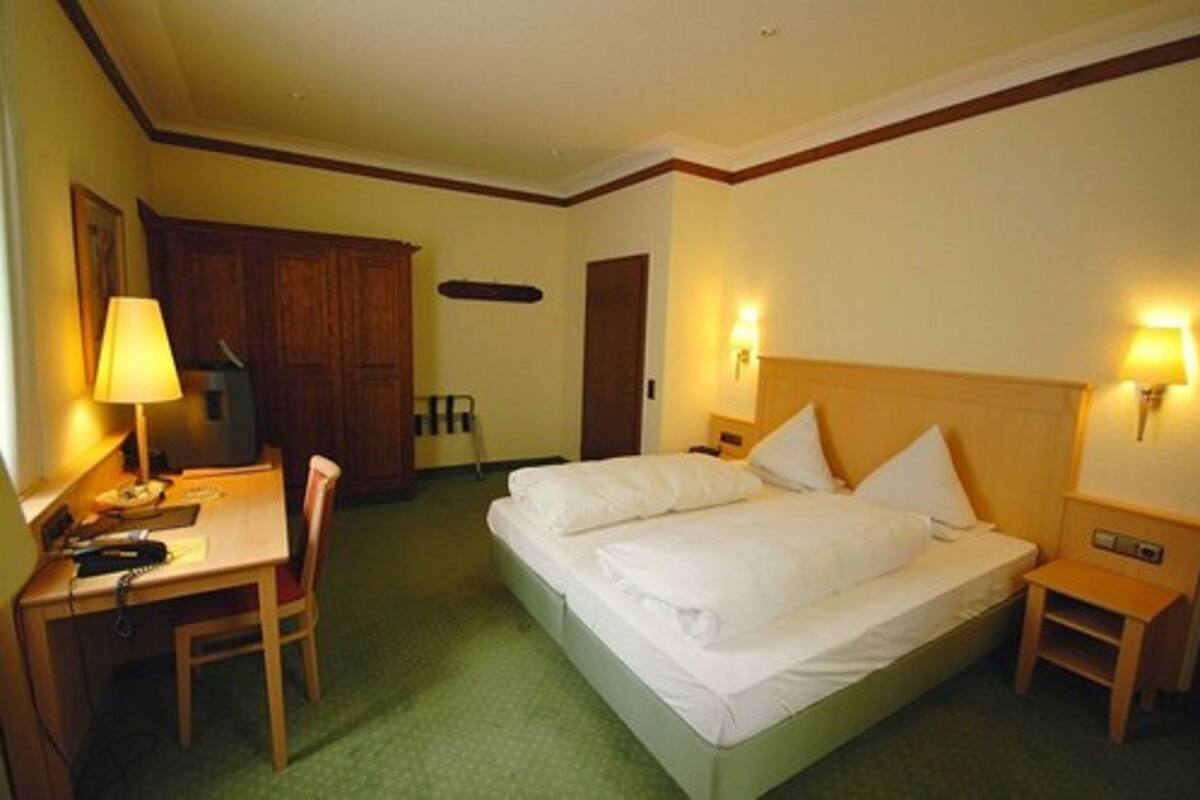 Quarto de hotel com cama, mesas de cabeceira, escrivaninha com cadeira, luminária e televisão.