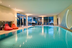 A modern indoor swimming pool with clear blue water, large windows, and a seating area with colorful bean bags. - Hotel Lamm (Heimbuchenthal)