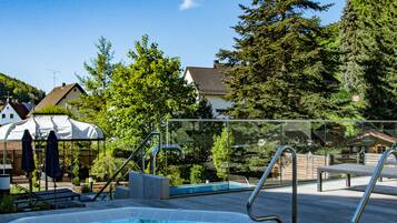 Un jacuzzi con cerramiento de vidrio, rodeado por una piscina, árboles y casas al fondo.