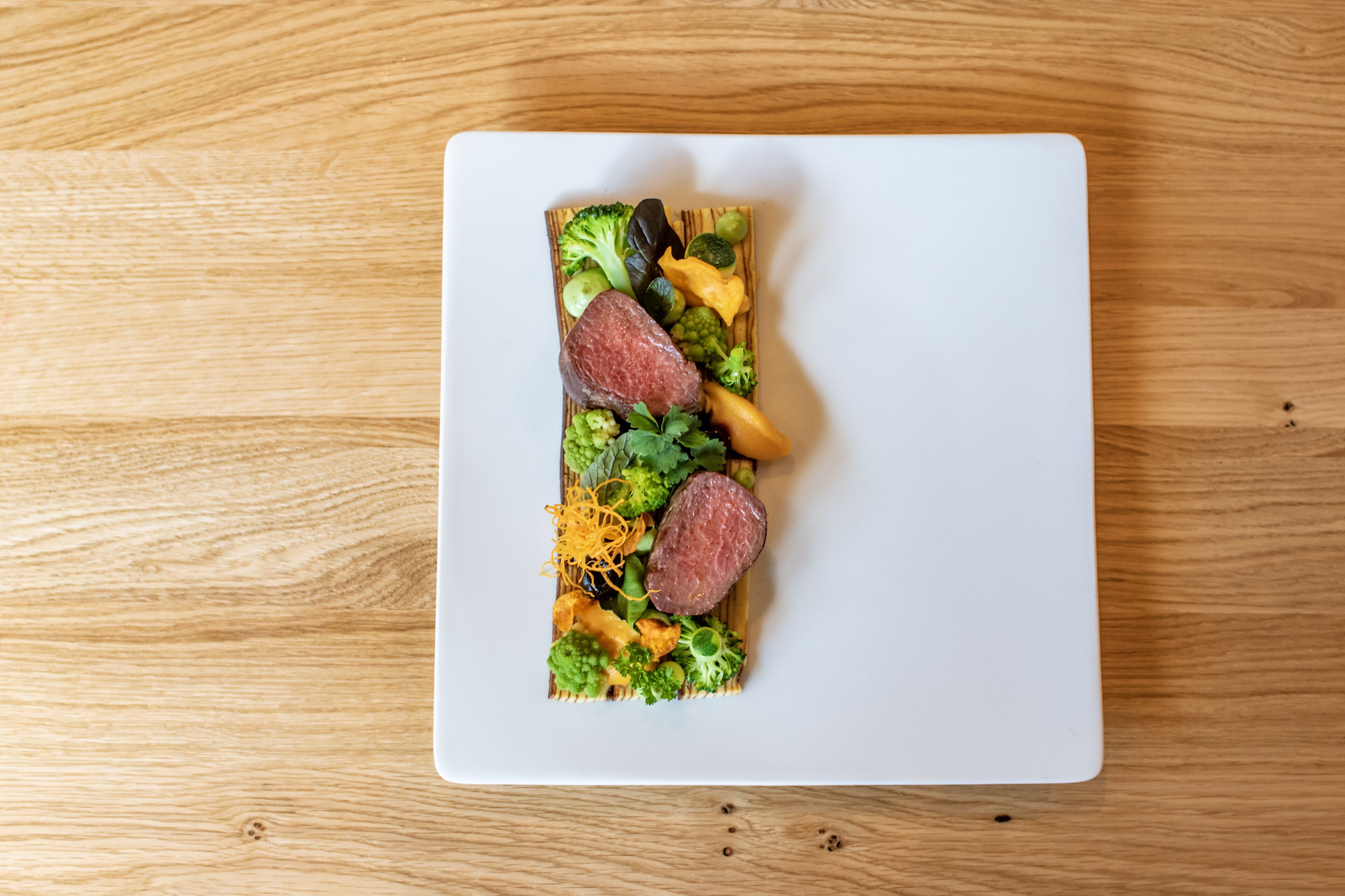 Ein weißer Teller mit verschiedenen Speisen, darunter Fleisch, Brokkoli und Käse, auf einem Holztisch.