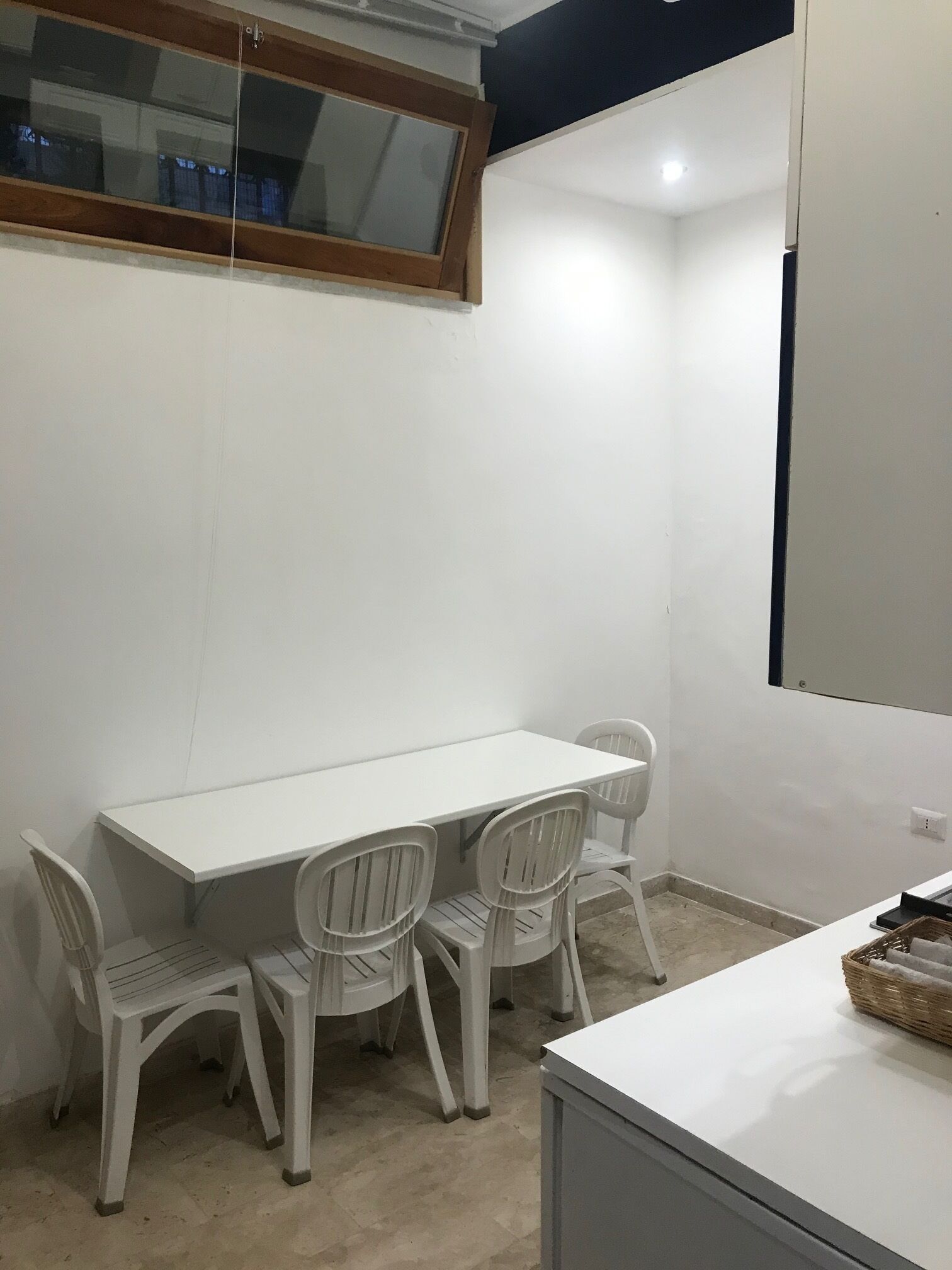 A small, clean room with a white table and four white chairs, a window with a wooden frame, and a white cabinet with a basket on top.