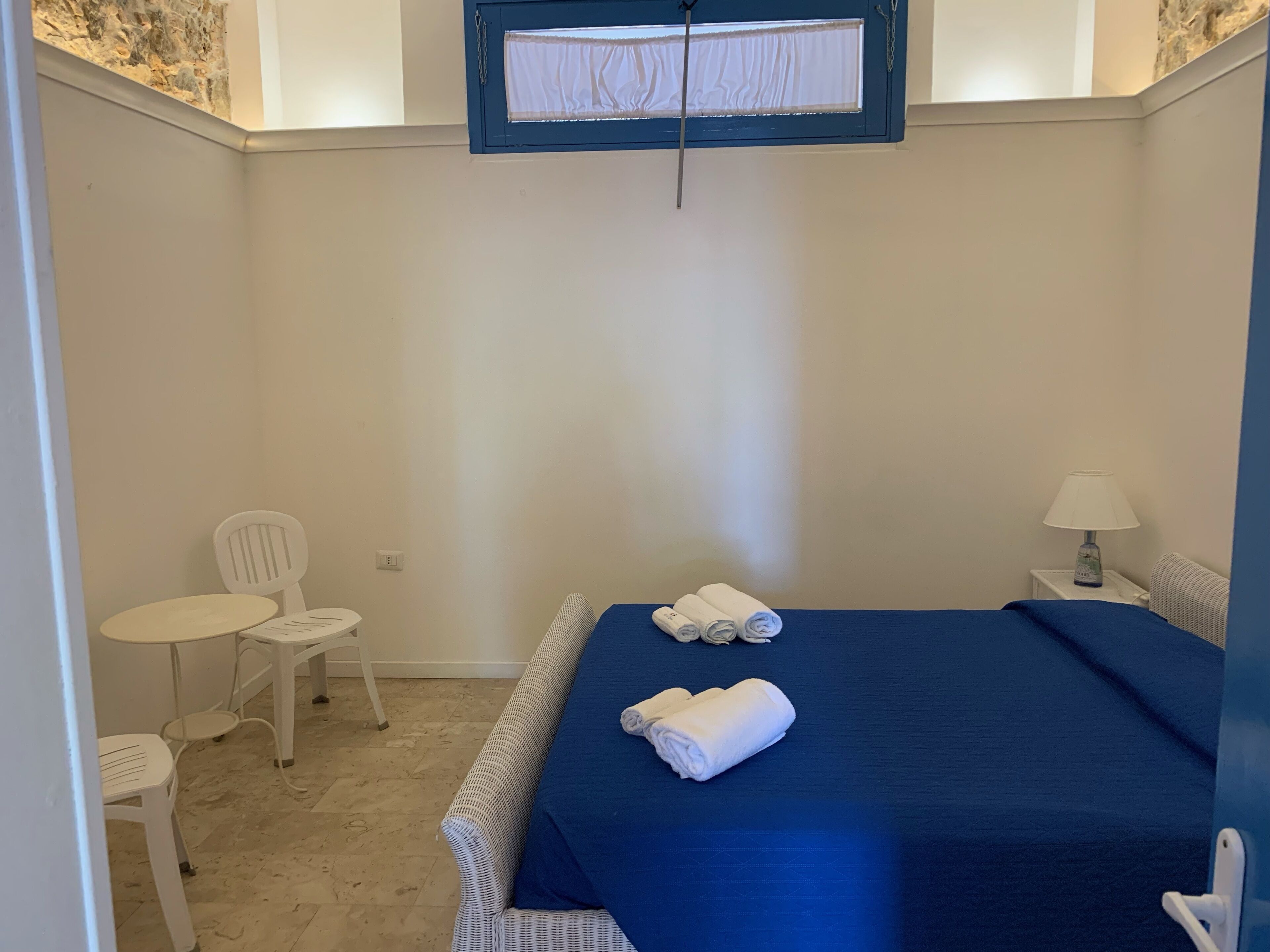 Una habitación pequeña y limpia con una cama, dos sillas, una mesita y un ventanal con cortina.
