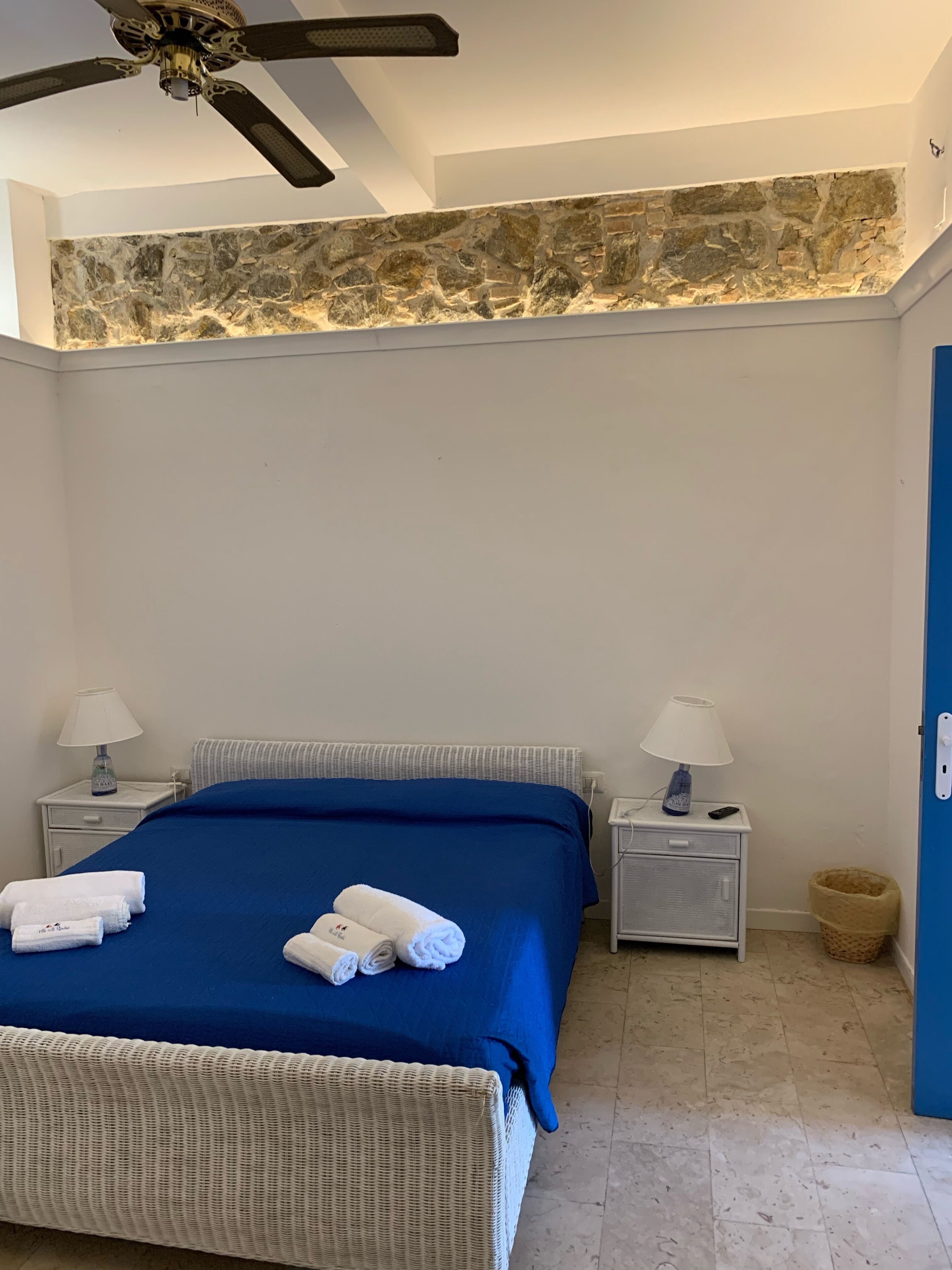 Une chambre à coucher avec un lit, deux tables de chevet avec des lampes, un ventilateur de plafond, un mur décoratif en pierre et une porte bleue.